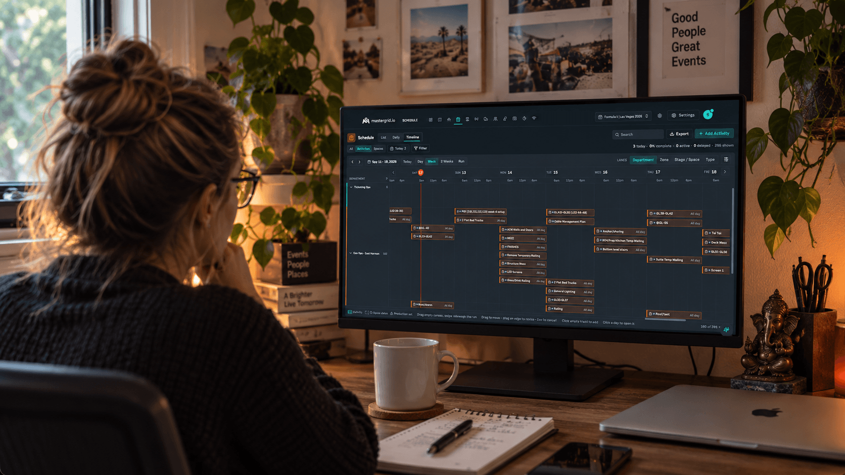 A production coordinator at a desk reading the MasterGrid Schedule timeline on a large monitor, a notebook and coffee in front of her.